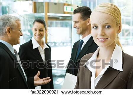 Young businesswoman smiling at camera, associates chatting in background View Large Photo Image Stock Image - Young businesswoman smiling at camera, associates chatting in background. Fotosearch