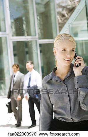 Young businesswoman using cell phone outdoors, looking away, businessmen walking in background View Large Photo Image Stock Image - Young businesswoman using cell phone outdoors, looking away, businessmen walking in background. Fotosearch