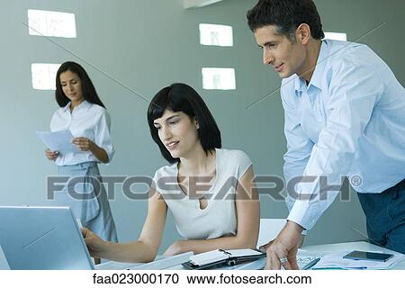 Young businesswoman working at computer, speaking to male colleague standing next to her View Large Photo Image Stock Image - Young businesswoman working at computer, speaking to male colleague standing next to her. Fotosearch