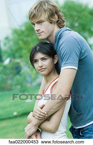 Stock Image - Young couple, man with arms around woman, woman leaning against him. Fotosearch