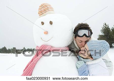 Stock Image - Young couple next to snowman, man kissing woman on cheek, eyes closed, portrait. Fotosearch