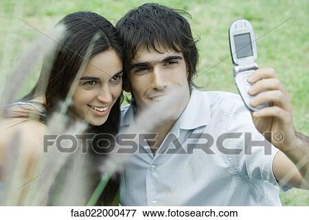 Young couple posing while man holds up cell phone to take photo View Large Photo Image Stock Photo - Young couple posing while man holds up cell phone to take photo. Fotosearch