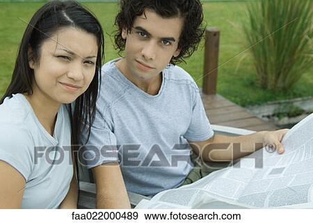 Young couple reading newspaper together outdoors, looking at camera View Large Photo Image Stock Photo - Young couple reading newspaper together outdoors, looking at camera. Fotosearch