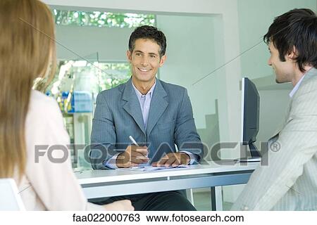 Stock Image - Young couple sitting across desk from smiling businessman, focus on businessman . Fotosearch