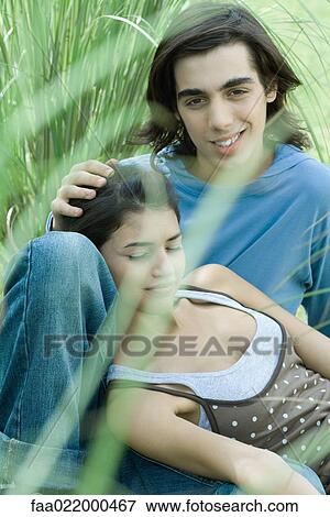 Stock Photo - Young couple sitting in grass, woman leaning head against boyfriend's knee. Fotosearch