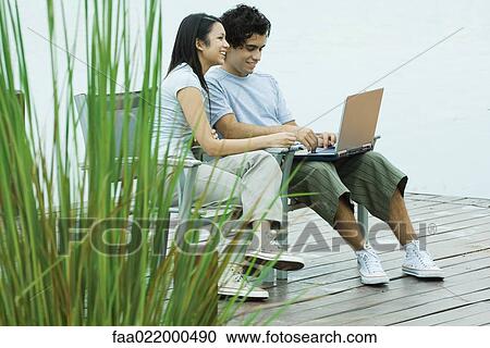 Stock Image - Young couple sitting on dock together, using laptop. Fotosearch