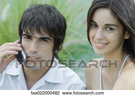 Young couple, woman looking at camera, man using cell phone, head and shoulders, portrait View Large Photo Image Stock Image - Young couple, woman looking at camera, man using cell phone, head and shoulders, portrait. Fotosearch