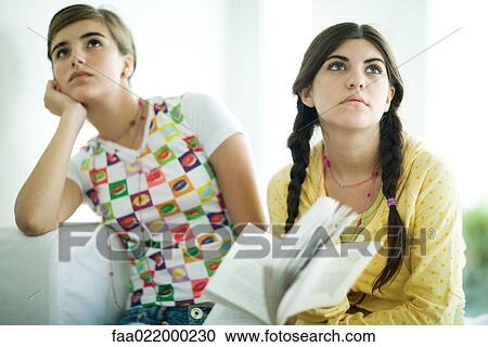Stock Image - Young female friends holding book together, looking away. Fotosearch