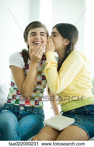 Stock Image - Young female friends, one whispering while the other laughs. Fotosearch