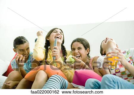 Young female friends sitting side by side, one singing into remote control as others laugh View Large Photo Image Stock Image - Young female friends sitting side by side, one singing into remote control as others laugh. Fotosearch