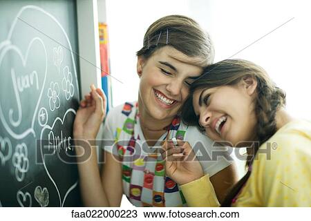 Stock Image - Young female friends writing names in hearts on chalkboard, laughing. Fotosearch