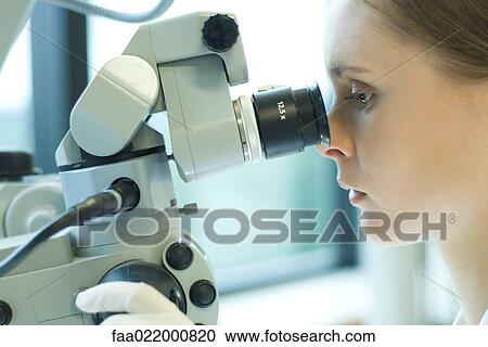 Stock Image - Young female scientist looking through microscope, extreme close-up. Fotosearch