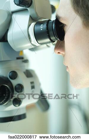 Stock Photo - Young male scientist looking through microscope, extreme close-up. Fotosearch