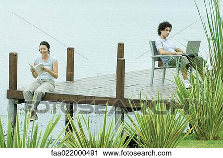 Young man and woman sitting apart on dock, woman reading book, man using laptop View Large Photo Image Stock Image - Young man and woman sitting apart on dock, woman reading book, man using laptop. Fotosearch