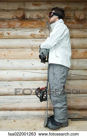 Stock Image - Young man holding snowboard, side view, full length. Fotosearch
