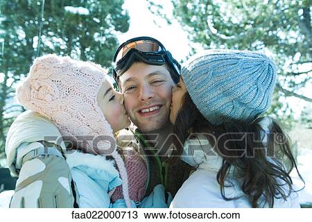 Stock Image - Young man in ski gear being kissed on cheeks by two young females. Fotosearch