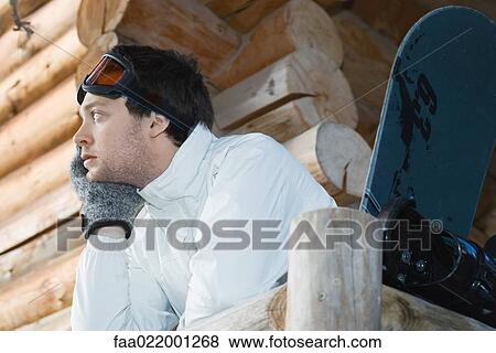 Stock Photo - Young man in ski gear standing against deck of log cabin, leaning on rail. Fotosearch