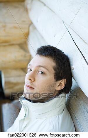 Stock Photograph - Young man leaning against wall of log cabin, looking at camera. Fotosearch