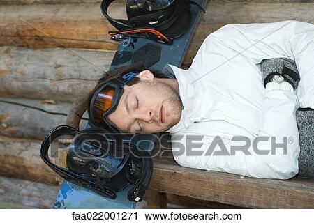 Stock Image - Young man lying on bench, resting head on snowboard, eyes closed. Fotosearch