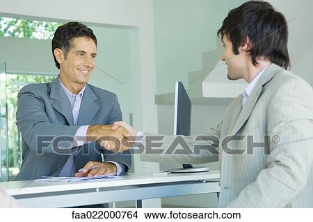 Picture - Young man shaking hands with mature businessman across desk, smiling. Fotosearch