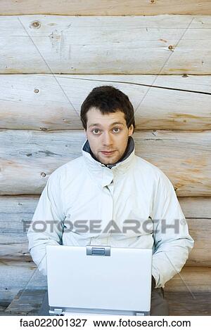 Stock Photo - Young man sitting next to wall of log cabin, using laptop. Fotosearch