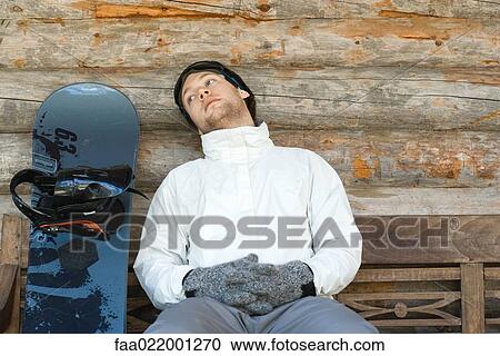 Stock Image - Young man sitting on bench next to snowboard, looking away. Fotosearch