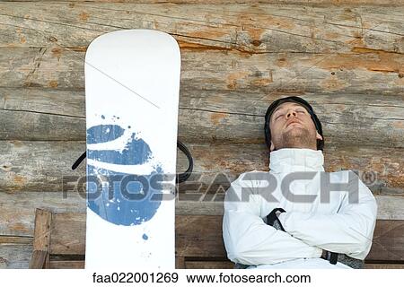 Stock Photo - Young man sitting on bench next to snowboard, napping. Fotosearch