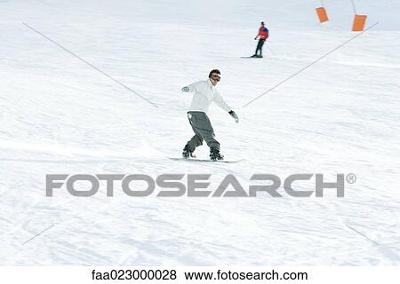 Young man snowboarding on ski slope, full length, skier in background View Large Photo Image Stock Photo - Young man snowboarding on ski slope, full length, skier in background. Fotosearch