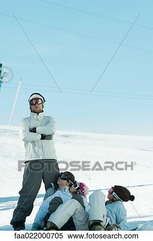 Young man standing with arms crossed in snow, young females on ground looking up at him View Large Photo Image Stock Photography - Young man standing with arms crossed in snow, young females on ground looking up at him. Fotosearch