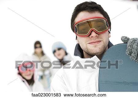 Stock Image - Young man standing with snowboard, friends in background, portrait. Fotosearch