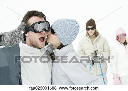 Stock Photo - Young man standing with snowboard, giving thumbs up sign as teen girl kisses his cheek. Fotosearch