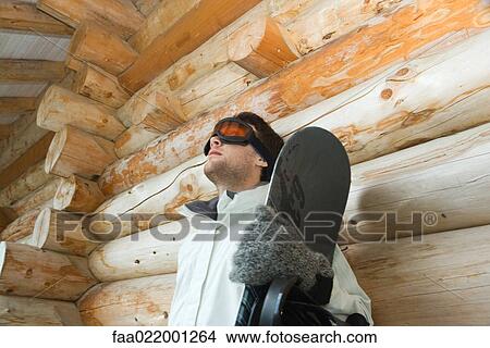 Picture - Young man with snowboard, leaning against log cabin, low angle view. Fotosearch