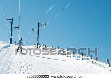 Stock Image - Young skier going up hill on ski lift, rear view. Fotosearch