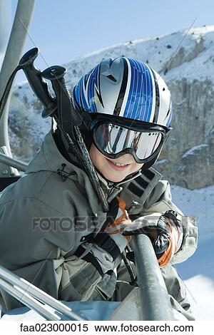Stock Photography - Young skier in chair lift, dressed in ski goggles and helmet, smiling at camera, portrait. Fotosearch