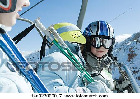 Stock Photo - Young skiers on chair lift, one whispering in the other's ear. Fotosearch