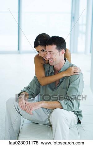 Stock Photo - Young woman embracing boyfriend from behind, man closing eyes, smiling. Fotosearch