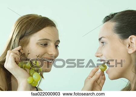 Stock Image - Young woman feeding her friend grapes, both smiling. Fotosearch
