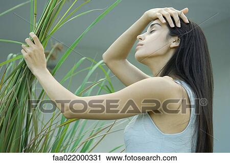 Stock Image - Young woman holding handful of plant stems, pushing hair back, eyes closed. Fotosearch