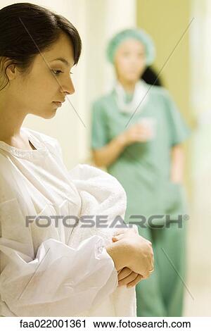 Stock Image - Young woman holding towel in hospital corridor, looking down, medical worker in background. Fotosearch