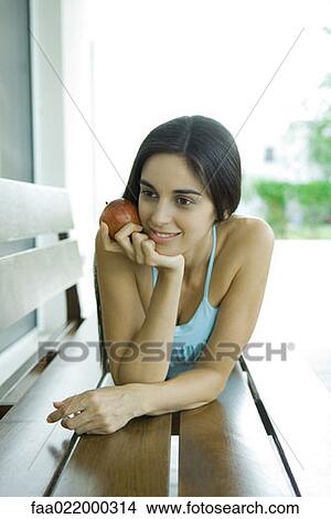 Picture - Young woman lying on bench, holding apple. Fotosearch