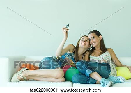 Young woman photographing self and friend with cell phone, sitting on couch View Large Photo Image Stock Image - Young woman photographing self and friend with cell phone, sitting on couch. Fotosearch