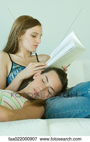 Stock Photo - Young woman reading book, friend resting head on her lap with eyes closed. Fotosearch