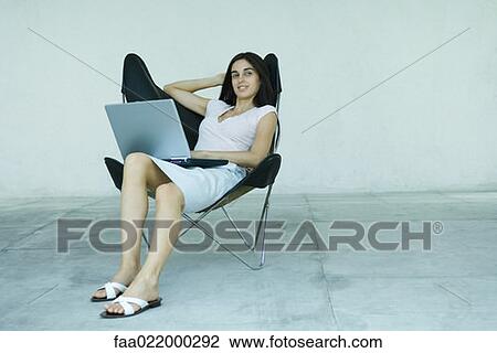 Young woman sitting back in chair, using laptop, full length portrait View Large Photo Image Stock Image - Young woman sitting back in chair, using laptop, full length portrait. Fotosearch
