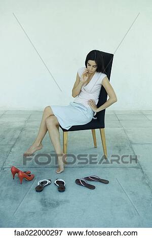 Stock Photo - Young woman sitting in chair, looking down at three pairs of shoes, full length portrait. Fotosearch