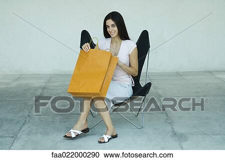 Stock Image - Young woman sitting in chair, opening shopping bag, smiling, full length portrait. Fotosearch