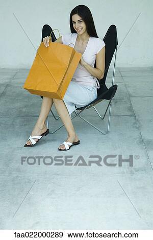 Young woman sitting in chair, opening shopping bag, smiling, full length portrait View Large Photo Image Stock Photo - Young woman sitting in chair, opening shopping bag, smiling, full length portrait. Fotosearch