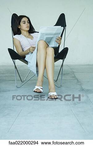 Young woman sitting in chair with legs crossed, reading newspaper, full length portrait View Large Photo Image Picture - Young woman sitting in chair with legs crossed, reading newspaper, full length portrait. Fotosearch