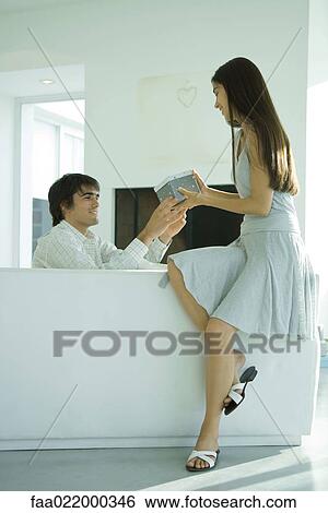 Young woman sitting on back of sofa, receiving gift from man, full length View Large Photo Image Stock Photograph - Young woman sitting on back of sofa, receiving gift from man, full length. Fotosearch