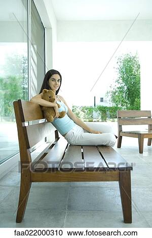 Stock Image - Young woman sitting on bench, holding teddy bear, full length portrait. Fotosearch