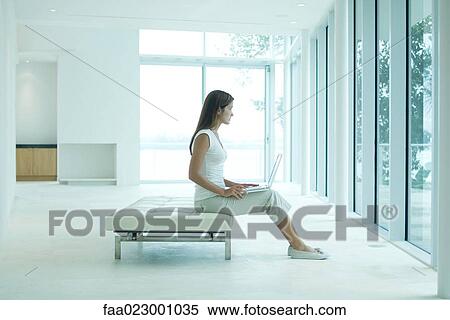 Stock Photography - Young woman sitting on settee, holding laptop computer, looking out window. Fotosearch